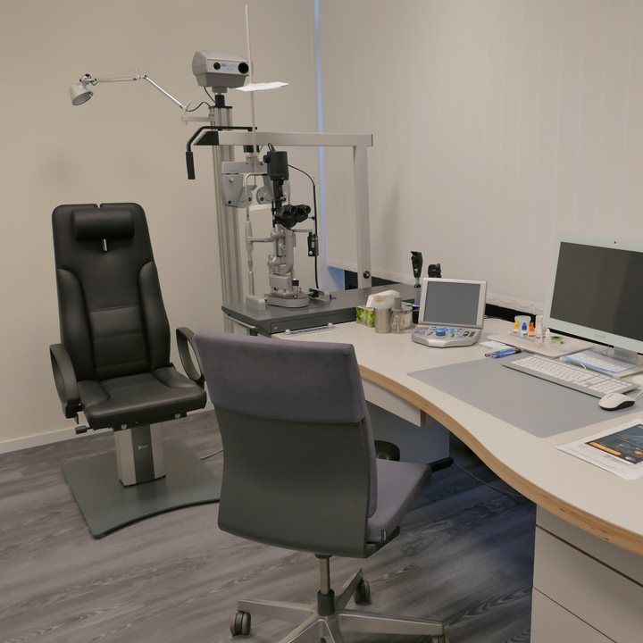 Behandlungsraum des Augenarztes. Man sieht ein Behandlungsstuhl und einen Schreibtisch mit Computer.